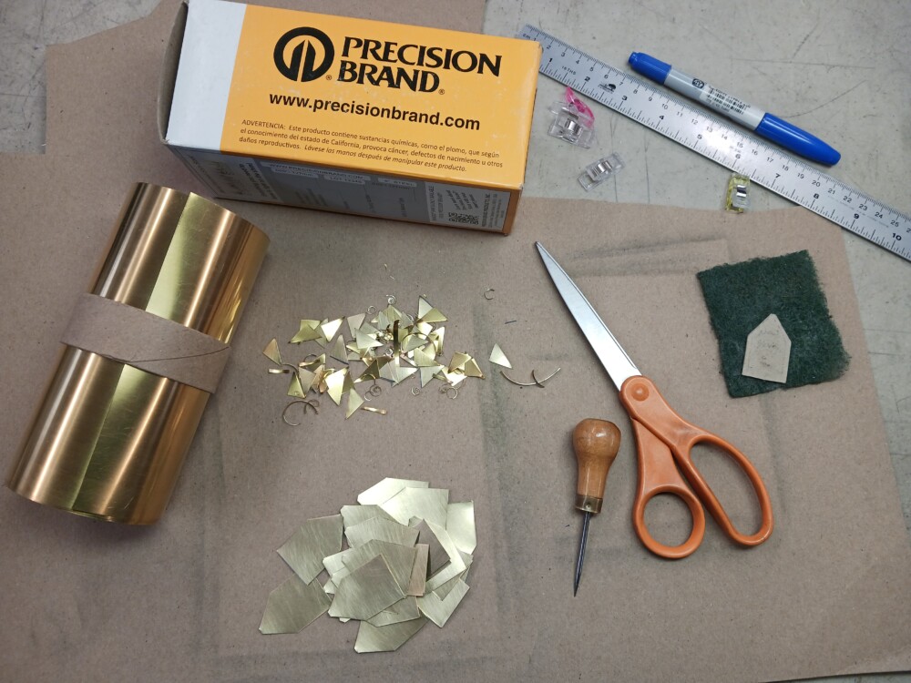 tools and materials for the project: roll of thin brass sheet, cut out backings, cut off scrap sheet, scissors, awl, green scrubby pad, marker, ruler, clips